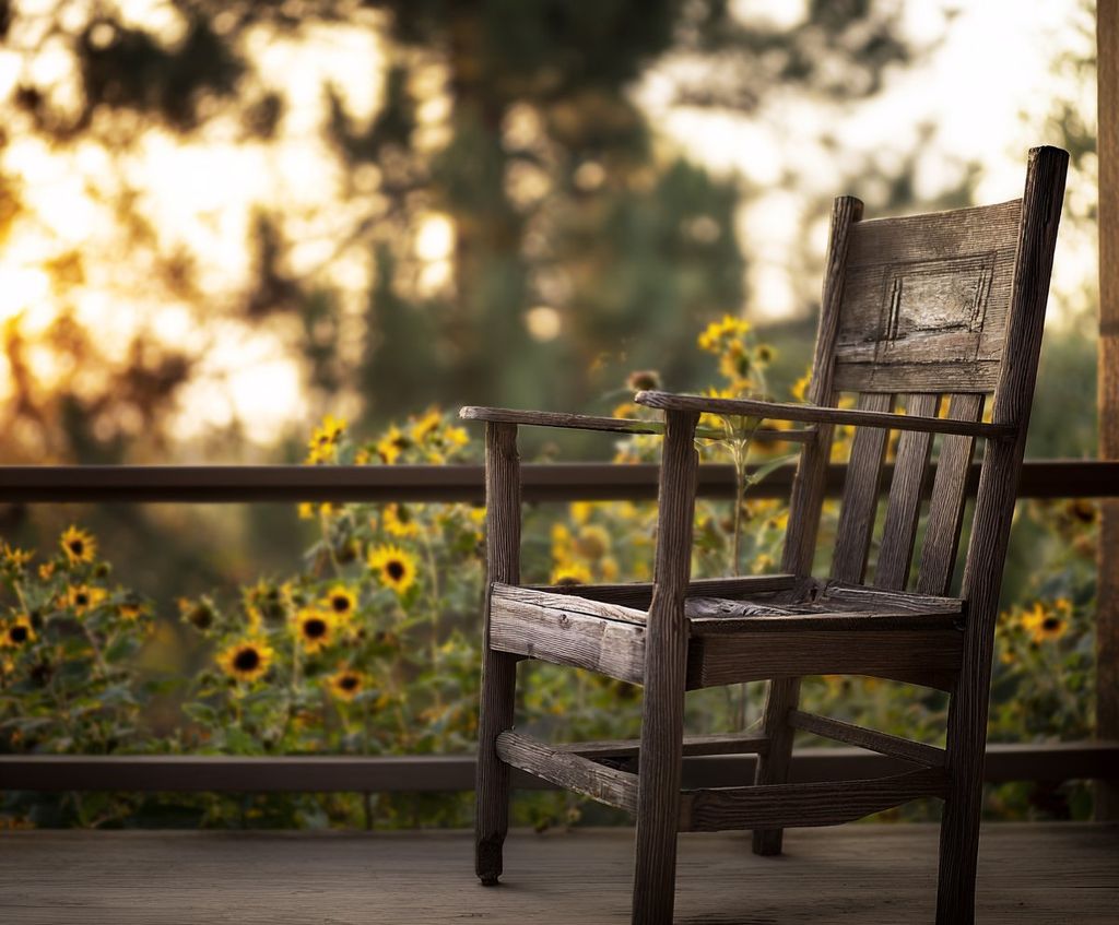 A weathered wooden chair on a porch with sunflowers beyond the rail